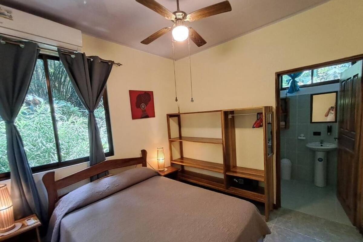 Ein Schlafzimmer mit einem Bett, einem Deckenventilator, einem Nachttisch mit Lampe, einem Fenster mit Vorhängen und Blick ins Freie.