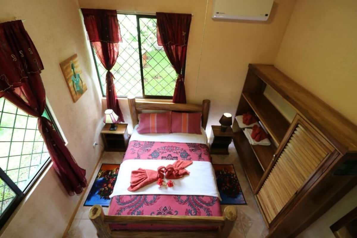 Ein kleines, gemütliches Zimmer mit einem Bett, einem Nachttisch, einem Holz-Kopfteil, einem Fenster mit Vorhängen und einem kleinen Tisch mit einer Lampe.