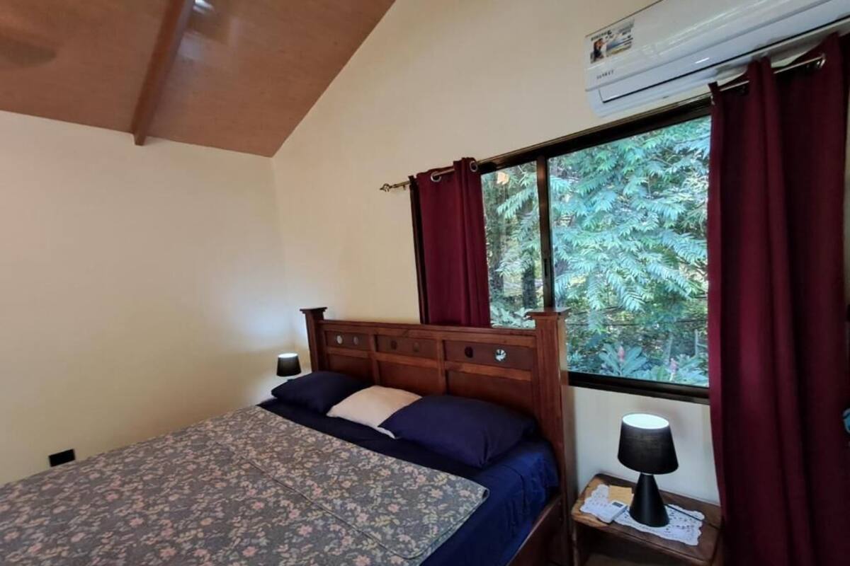 Ein Schlafzimmer mit Bett, Nachttisch, Lampe, einem Fenster mit Vorhängen und einer Klimaanlage.