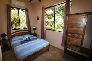 Ein Schlafzimmer mit Bett, Nachttisch, Lampe, Kleiderschrank und einem Fenster mit Blick ins Grüne.