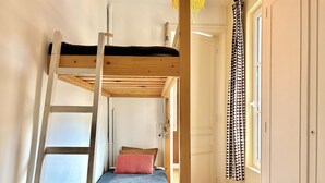 Uma cama beliche com escada, uma cama de solteiro com travesseiro vermelho, piso de madeira, uma porta branca e uma janela com cortinas estampadas.