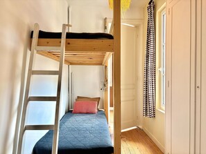 Ein Etagenbett mit einer Leiter, ein Einzelbett mit einem roten Kissen, Holzboden, eine weiße Tür und ein Fenster mit gemusterten Vorhängen.