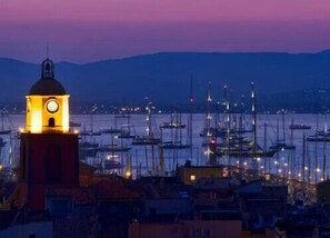 Eine Küstenstadt im Abendlicht mit einem markanten Glockenturm, einem Hafen voller Boote und einem Berg im Hintergrund.