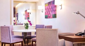 Habitación de hotel con una mesa redonda, sillas moradas, un escritorio con teléfono, una lámpara y un espejo grande.