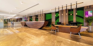 Un área de recepción moderna con un mostrador de madera, dos sillas, una planta grande en maceta y una pared verde con listones de madera verticales.