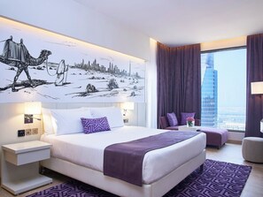 Una habitación de hotel moderna con una cama grande, un mural con un paisaje urbano, vistas de un edificio alto y una zona de estar con una alfombra morada.