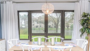Un comedor con mesa de madera, sillas blancas, una lámpara colgante y vistas a través de puertas de cristal.