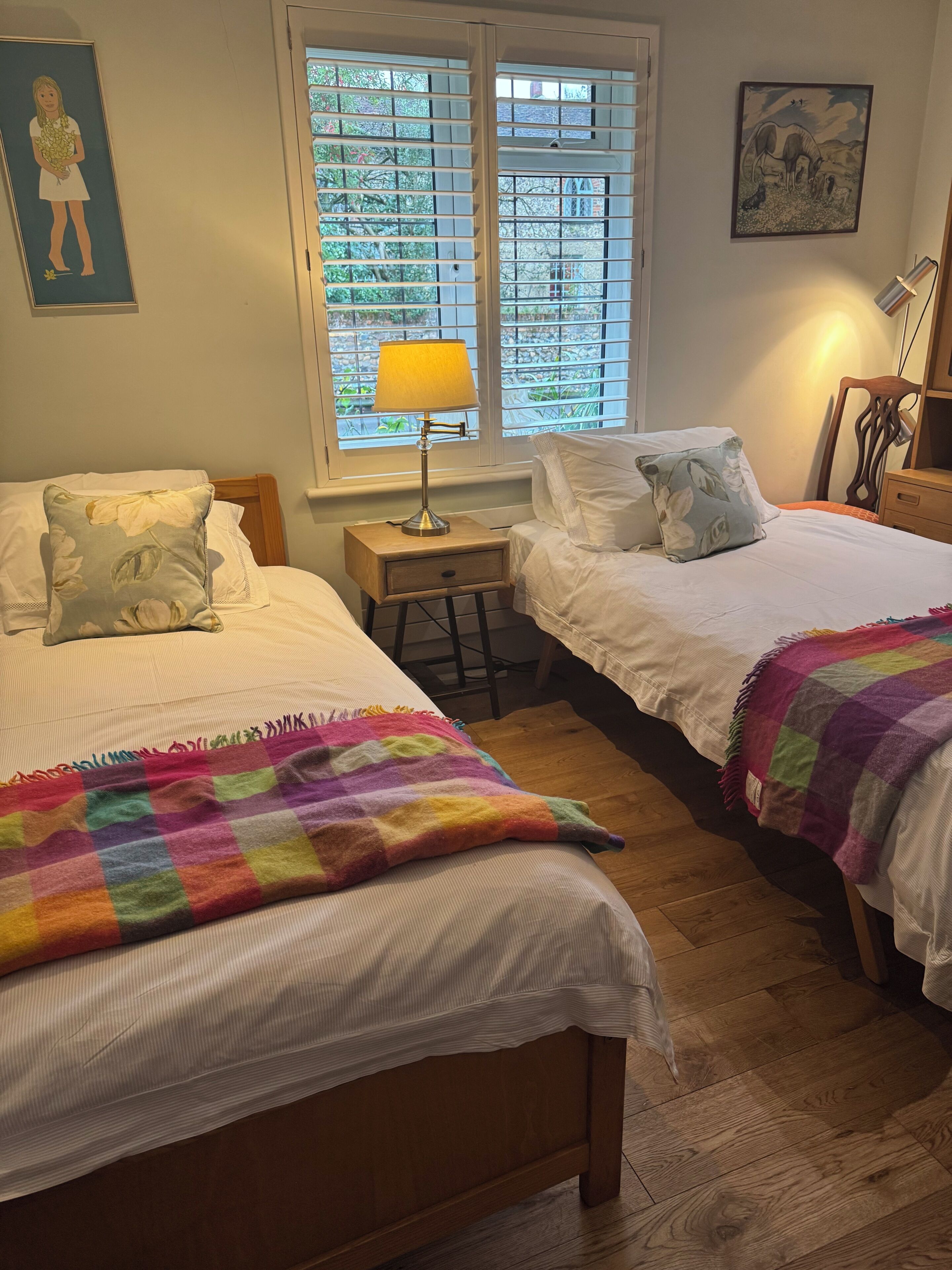 Un dormitorio con dos camas, una ventana con persianas, una lámpara de noche y una manta colorida.