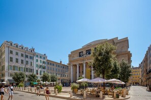 Piazza con tavoli all'aperto, un edificio imponente con colonne e cielo azzurro intenso.