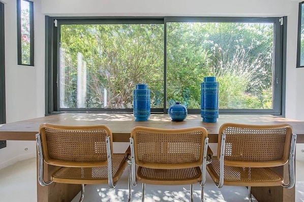 Ein Essbereich mit einem Tisch, vier Rattan-Stühlen, zwei blauen Vasen und Blick auf Grün durch große Fenster.