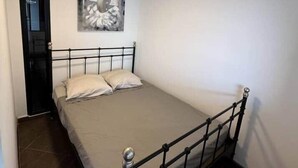 Yhden hengen sänky, jossa on metallirunko, beige patja ja kaksi tyynyä. Vasemmalla puolella on ovi ja sängyn yläpuolella seinälle ripustettu taideteos.