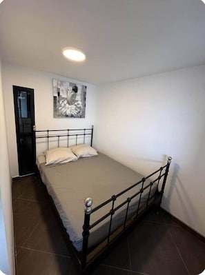 A single bed with a metal frame, a beige mattress, and two pillows. There is a door to the left and a wall-mounted artwork above the bed. - 1-bedroom apartment on the Corniche (Marseille)