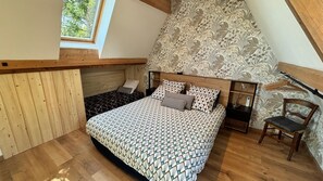 Quarto com teto inclinado, cama com roupa de cama estampada, uma cadeira e uma mesa de cabeceira.