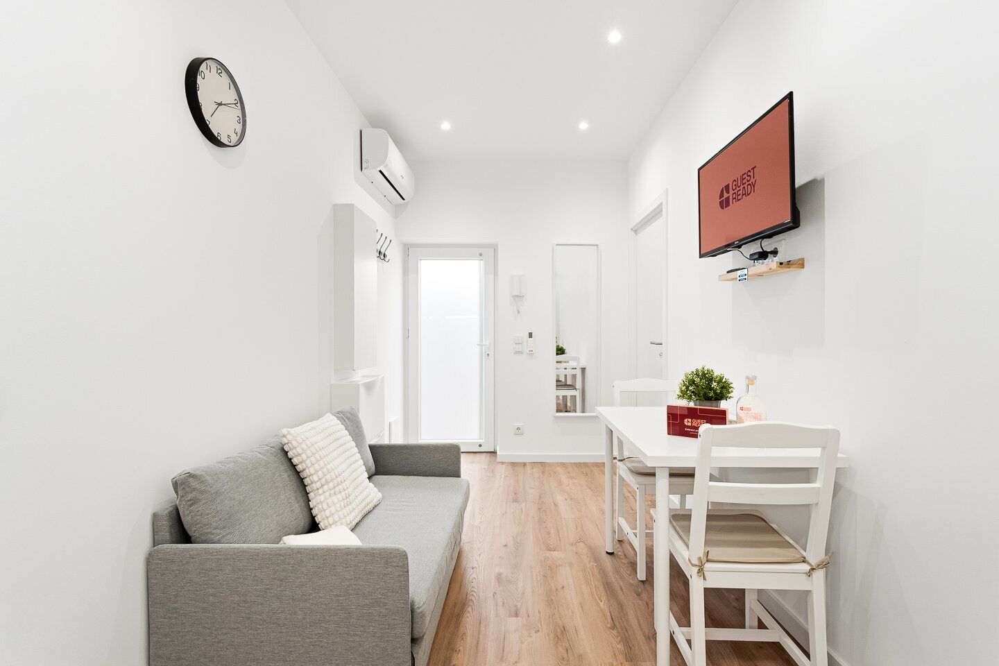 Ein moderner, minimalistischer Wohnraum mit einem grauen Sofa, einem Esstisch mit Stühlen, einem Flachbildfernseher und einer Wanduhr.