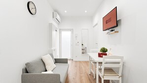 Et moderne, minimalistisk opholdsrum med en grå sofa, et spisebord med stole, et fladskærms-tv og et vægmonteret ur.