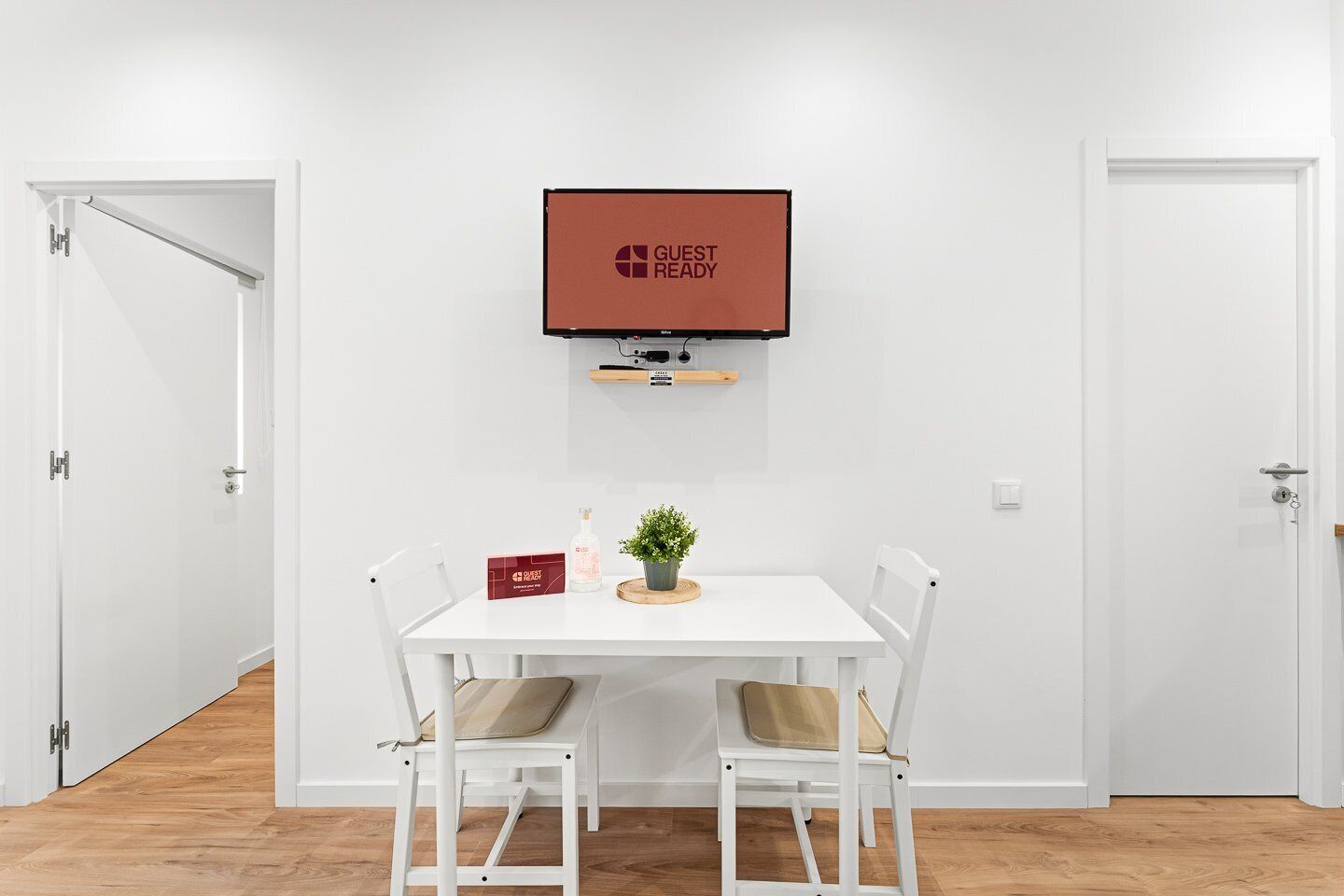 Une pièce avec une table blanche, deux chaises, une petite plante, une bouteille et une boîte rouge. Un téléviseur est fixé au mur, au-dessus de la table.