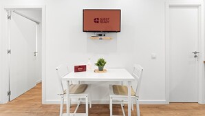 Et værelse med et hvidt bord, to stole, en lille plante, en flaske og en rød æske. Et fjernsyn er monteret på væggen over bordet.