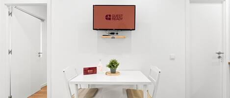 Una habitación con una mesa blanca, dos sillas, una planta pequeña, una botella y una caja roja. Un televisor está montado en la pared sobre la mesa.