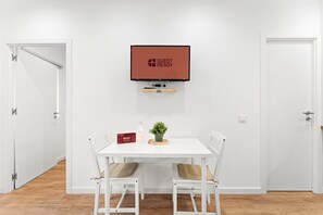 Ein Zimmer mit einem weißen Tisch, zwei Stühlen, einer kleinen Pflanze, einer Flasche und einer roten Kiste. Über dem Tisch ist ein Fernseher an der Wand befestigt.