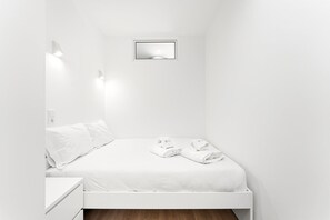 Pieni, valkoinen huone, jossa on yksi sänky, yöpöytä, seinään kiinnitetty valaisin ja pieni ikkuna.