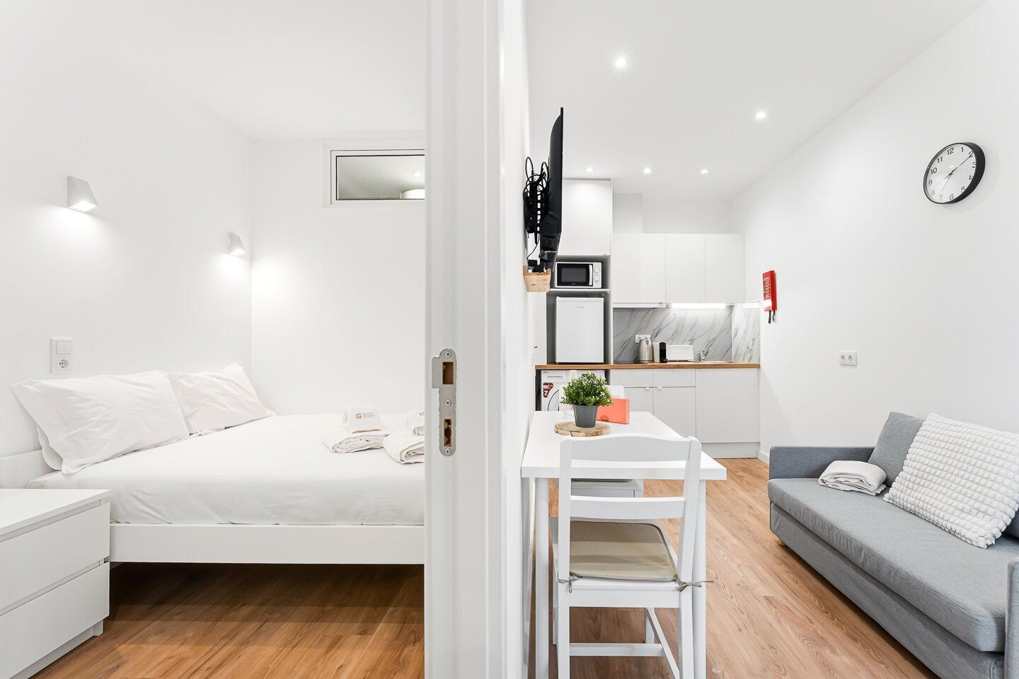 A modern, white-themed room with a bed, a small dining table with a chair, a grey sofa, and a kitchen area with a microwave and a clock on the wall.