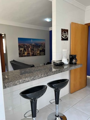 A kitchen with a granite countertop, two black bar stools, a coffee maker, and a painting of a cityscape. - LK Santo Antônio 1 (Belo Horizonte)