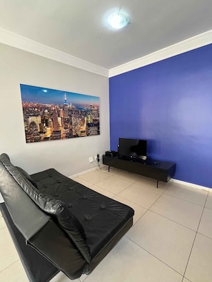 A room with a black sofa, a flat-screen TV on a black stand, a cityscape painting on the wall, and a blue accent wall. - LK Santo Antônio 1 (Belo Horizonte)
