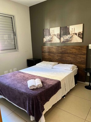 Un dormitorio con cama, cabecera, ventana y dos cuadros colgados en la pared.