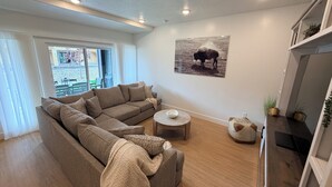 Una sala de estar con un sofá modular, una mesa de centro redonda, un cuadro de un bisonte y un ventanal corredizo que da a un espacio exterior.
