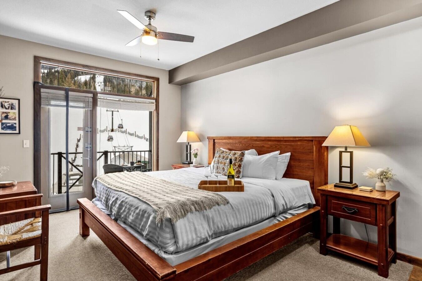 Une chambre avec un lit en bois, deux lampes de chevet, un ventilateur de plafond, une table de chevet en bois et une vue sur un balcon extérieur, accessible par une porte vitrée.