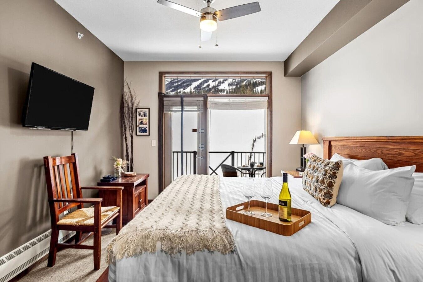 A hotel room with a bed, a desk, a chair, a TV, a balcony with a view, and a ceiling fan.