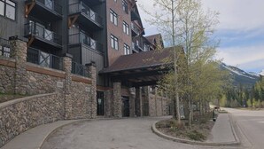 A multi-story building with stone and brick exterior, a covered entrance, and a mountain backdrop. - Golden Alpine Hideaway Studio Hot tub + Ski-in/out (Golden)