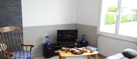 Una sala de estar con una mecedora de madera, un sofá gris, una mesa de centro con una bandeja de comida, un televisor y una ventana con vistas a la vegetación.