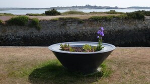 Una jardinera grande con plantas y una flor morada en primer plano, un muro de piedra, y vista al mar y tierra a lo lejos.