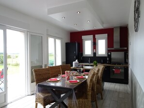 A dining area with a table set for four, a kitchen with modern appliances, and a sliding glass door leading to a balcony. - Holiday Home Near the Beach (Saint-Marcouf)