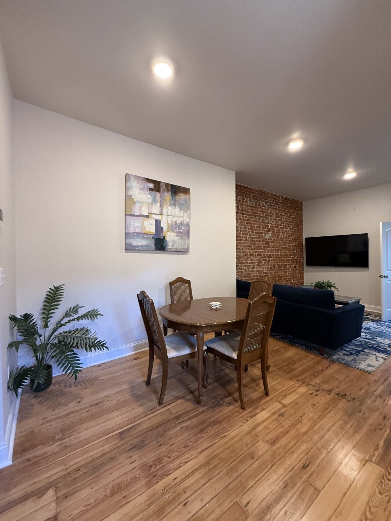 Ein Essbereich mit Holztisch und -stühlen, einem Bild an der Wand, einer Wand mit Backsteinoptik, einem an der Wand befestigten Fernseher und einem blauen Sofa im Wohnbereich.