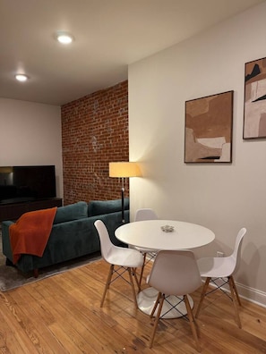 A living room with a brick wall, a sofa, a TV, a round table with chairs, and two abstract paintings. - Benton Park Corner- IN2S (St. Louis)