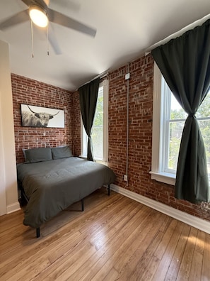 A bedroom with a brick wall, a bed, a ceiling fan, and a window with curtains. - Benton Park Corner- IN2S (St. Louis)
