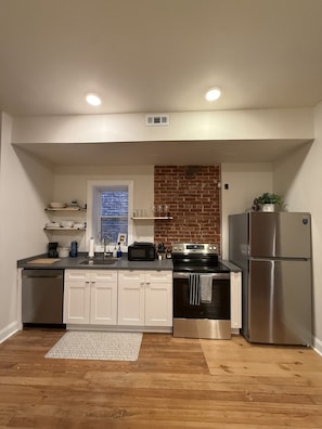 A modern kitchen with white cabinets, a brick accent wall, a stainless steel refrigerator, and a window with a view. - Benton Park Corner- IN2S (St. Louis)