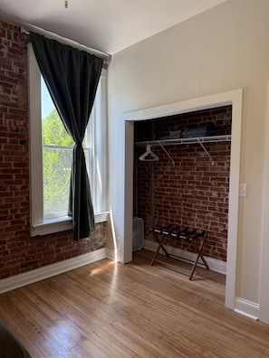 A room with a brick wall, wooden flooring, a window with curtains, a fan, and a metal rack with hangers. - Benton Park Corner- IN2S (St. Louis)