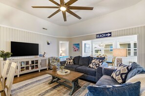 Een woonkamer met een flatscreen televisie, een plafondventilator, een bank, een salontafel en een bord dat de richting naar het strand aangeeft.