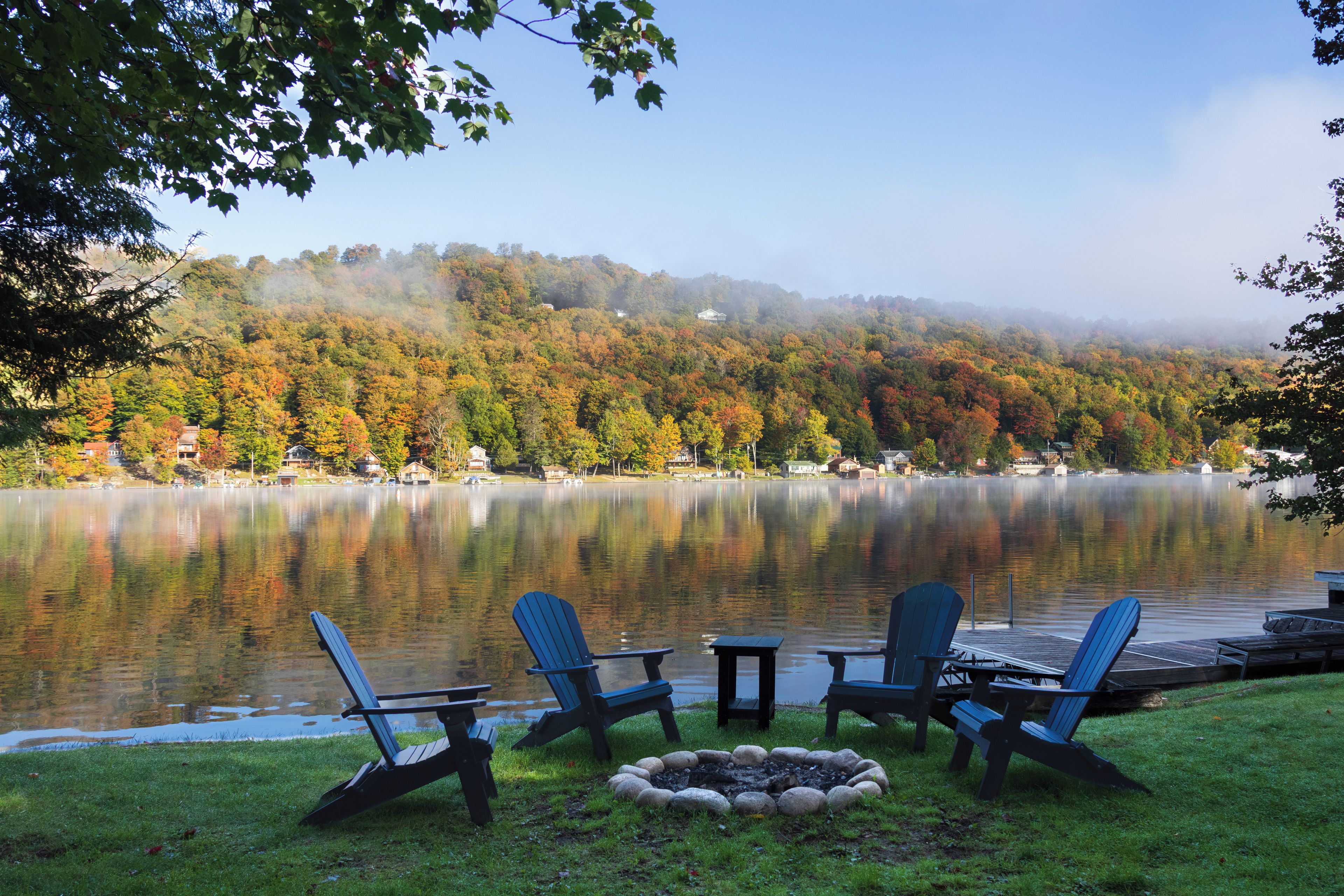 A lakeside view with Adirondack chairs, a fire pit, and a dock extending into the water.
