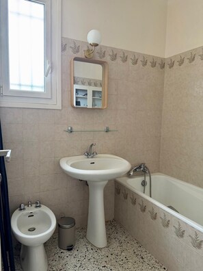 Ein Badezimmer mit einem Standwaschbecken, einem Bidet, einer Badewanne, einem Spiegel mit Wandleuchte und einem Fenster.