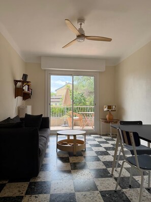 Ein Wohnzimmer mit schwarzem Sofa, rundem Couchtisch, Esstisch mit Stühlen, Deckenventilator und Blick ins Freie durch eine Glastür.