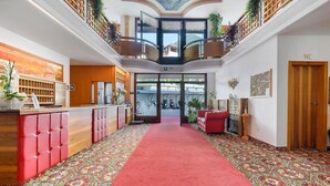 A hotel lobby with a red carpet, a reception desk, a red sofa, and a balcony with plants. - Das Bergland – Jaufen Apartment with Covered Panoramic Terrace and Mountain Views (St. Leonhard in Passeier)