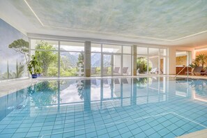 Indendørs swimmingpool med glasvægge, liggestole og udsigt til bjerge.