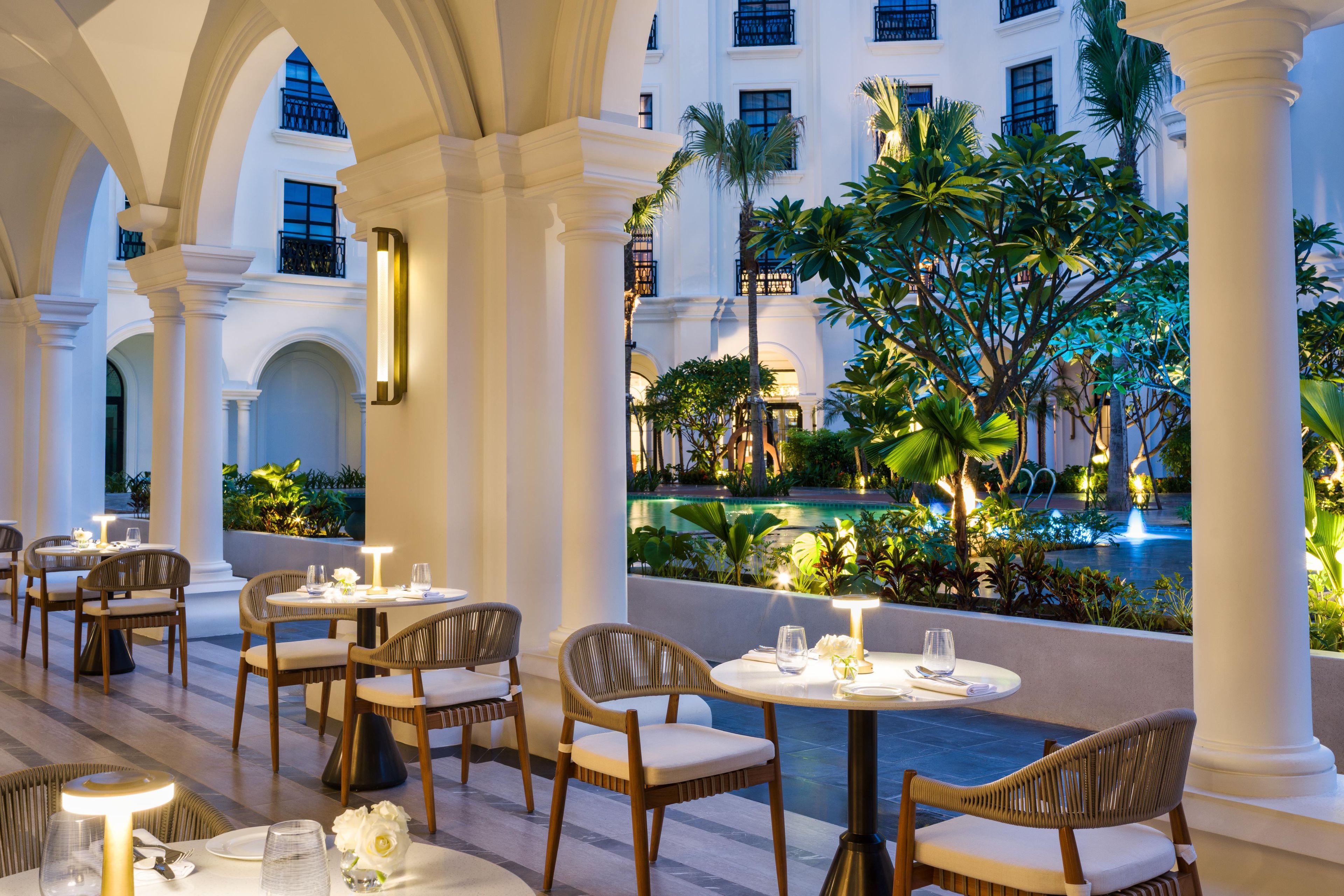 A restaurant with outdoor seating, white columns, and a view of a pool and greenery.
