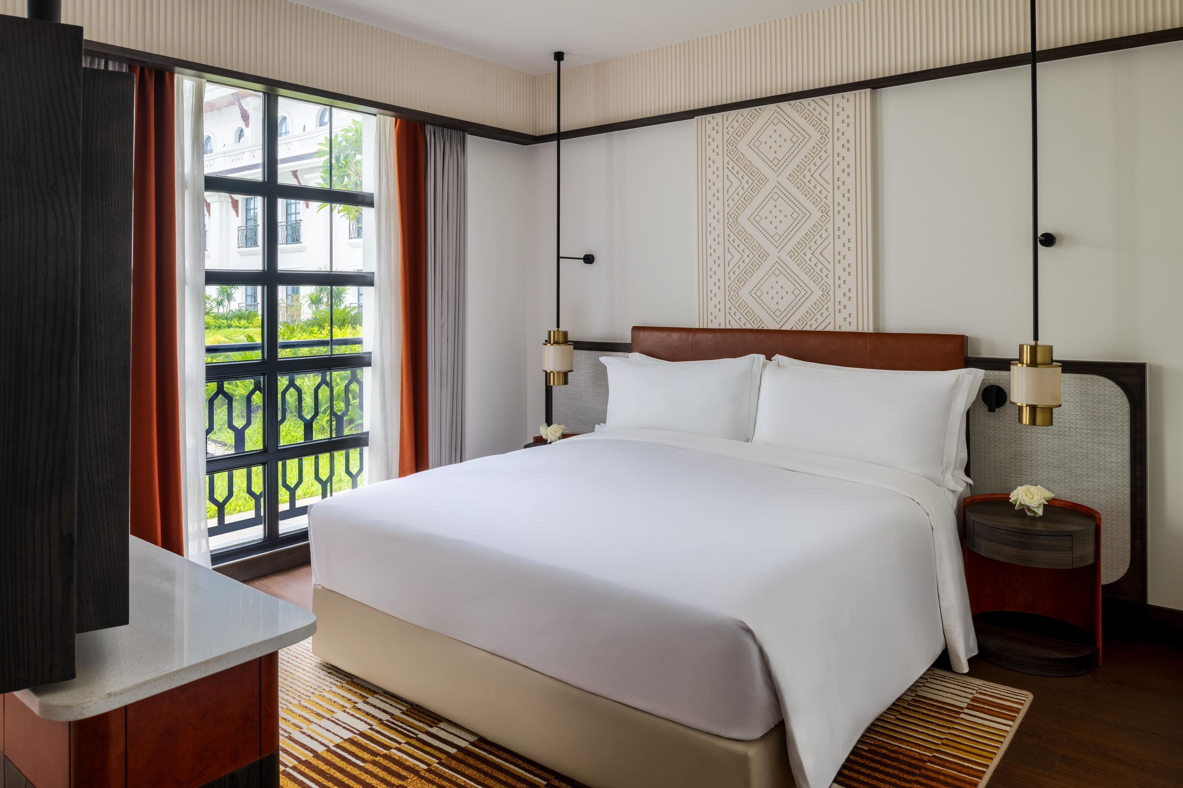 Une chambre d’hôtel avec un grand lit, une table de chevet, une commode, une fenêtre donnant sur un paysage verdoyant et un mur décoré de motifs.