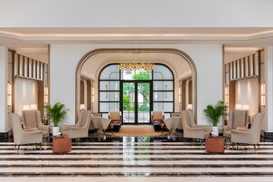 A hotel lobby with a grand arched entrance, marble flooring, and seating areas with lamps and potted plants.