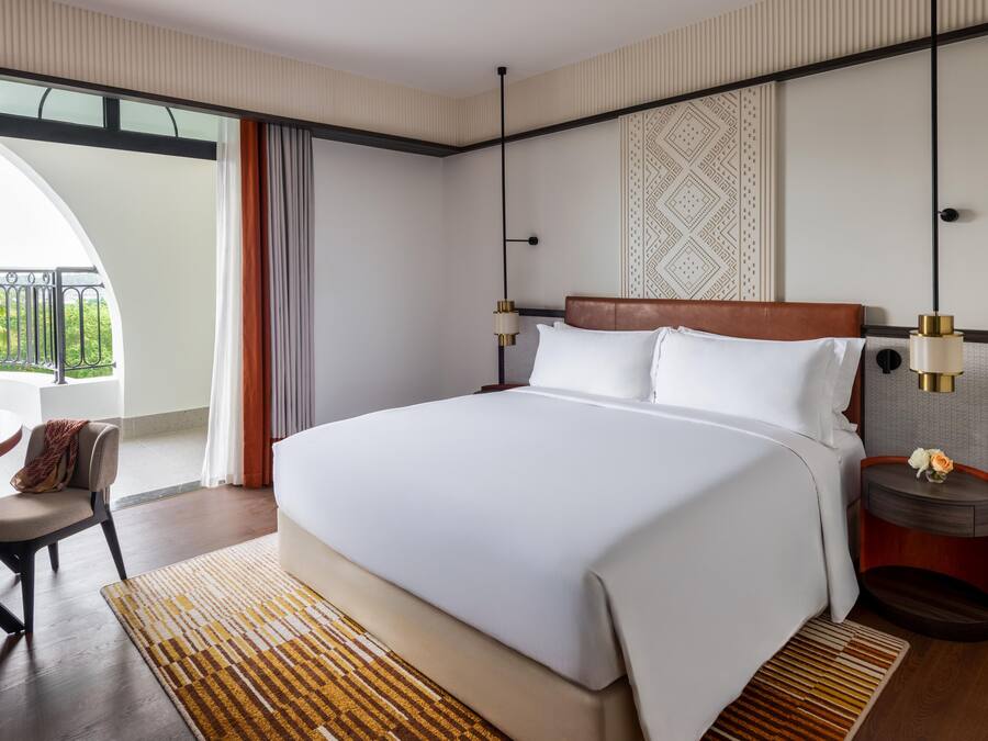 A hotel room with a large bed, a desk, a chair, a bedside table with a lamp, and a view of greenery through the window.